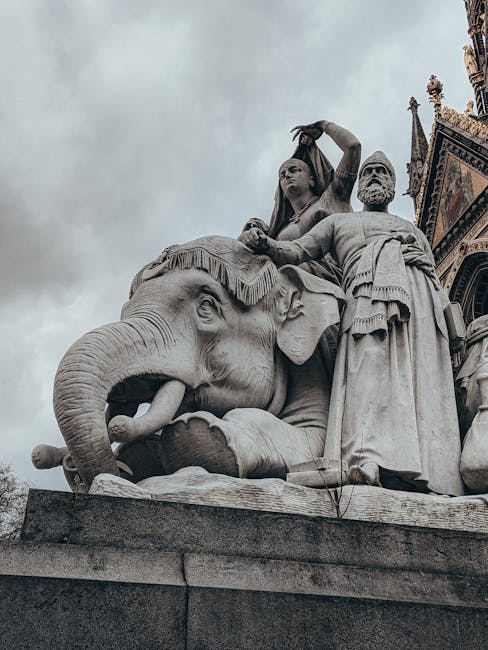 A detailed close-up of a large stone monument featuring a carved elephant with its trunk curled upward and detailed facial features, situated at the base of a historic building with ornate architectural elements partially visible on the right side. The statue includes a seated woman on top of the elephant holding a decorative object, and a standing man in classical attire beside the elephant, both with carved drapery and expressions. The monument appears to be made of light grey stone set against a cloudy sky background, emphasizing the intricate sculptural details and proportions. This outdoor sculpture is part of the architectural embellishments typical of historic European structures, often associated with cultural or historical significance, and may be located in a city environment where house removals or relocation services such as those offered by elephantandcastlemanandvan.co.uk are relevant for preserving or transporting similar artifacts during moving projects.