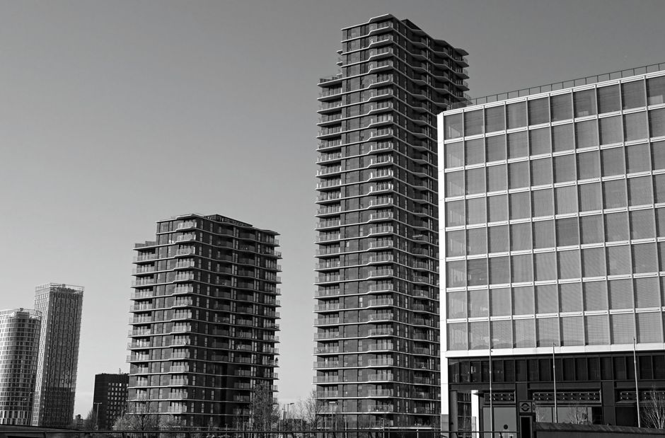 Black and white photograph of three modern high-rise buildings in an urban area, with two taller residential towers on the left and center, and an office building with a grid-like facade on the right.
