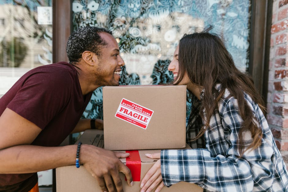 A man and a woman are engaged in a moving activity outside a building, with the man leaning forward and smiling at the woman, who is also smiling back. They are holding a cardboard box with a red and 