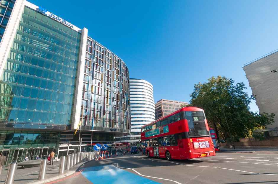 A bright red double-decker bus on a busy city street in Elephant and Castle, London, with modern office buildings featuring glass facades and a curved white office tower in the background. The street is lined with trees and a few pedestrians, with blue sky overhead. Nearby, a property entrance is visible, with part of a building’s doorway and pavement seen on the right side. The scene captures city traffic and urban architecture, relevant to home relocation and furniture transport services offered by companies like Elephant and Castle Man and Van. The image reflects typical moving logistics in a metropolitan environment, highlighting the importance of efficient loading and transportation during house removals or packing processes.