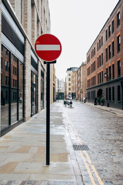 A narrow city street with a large red and white 'no entry' traffic sign mounted on a black pole positioned on the pavement. On the left side, there are multi-storey brick buildings with large glass windows, suggesting commercial or office spaces, while the right side features darker brick facades of similarly styled buildings. The street is paved with cobblestones, and the sidewalk is made of rectangular stone slabs. In the background, there are a few pedestrians and cyclists, with some pushed bicycles visible near the buildings. The scene appears to be in a quiet urban area potentially suitable for home removals or furniture transport, with no vehicles present on the road, which may indicate restrictions or a one-way system. This setting reflects an environment where careful logistical planning is necessary for house relocation or moving services, possibly involving navigating narrow access points for larger items, as seen in the context of Elephant and Castle removals for properties on Walworth Road.